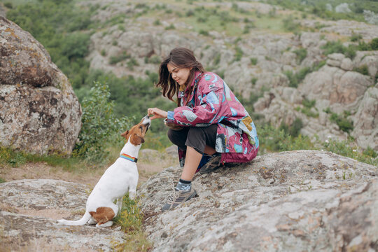 girl traveler in stylish clothes sits on a stone and feeds from the hands of a dog breed Jack Russell Terrier on a background of mountains