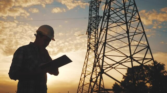 electrical engineer worker in helmet silhouette working at sunset near the tower with electricity. business energy technology industry concept. electrician studying reading documents on a tablet