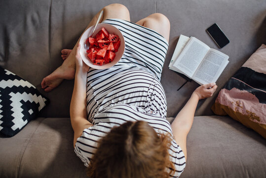 Pregnant woman reading a book while eating watermelon on a cozy couch