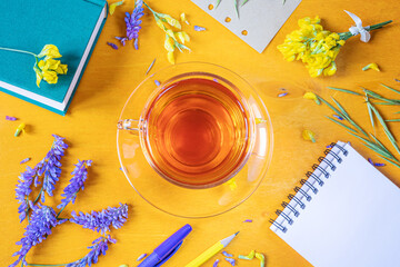 Cup of tea in a transparent cup with a saucer, notebook, pen, pencil, wildflowers and herbs on a yellow wooden background. Still life in yellow and purple tones. Summer cute funny flat lay.