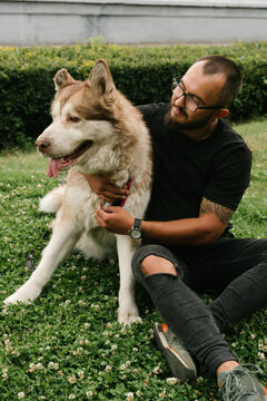 Short-haired guy with beard and glasses with the malamut dog sit on the lawn at the park