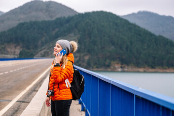 Side view of a woman in orange coat and wool hat using a mobile phone on a road with mountains in the background