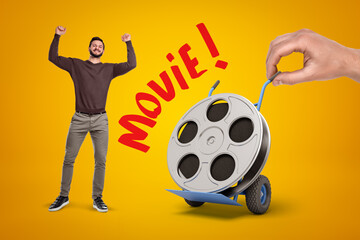 Happy man in casual clothes and hand holding hand truck with film reel and 'Movie' sign on yellow background.