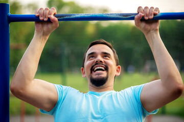 Naklejka premium Cheerful middle-aged man doing pull ups exercise on shabby steel bar outdoors in summer evening. Tanned bearded man in blue shirt doing workout at training ground. Strength and motivation concept