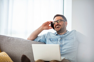 Young man sitting relaxed on sofa with laptop checking his smartphone messages. Laptop and smartphone mockup - young man with computer and phone sitting
