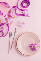 Flower and table settings overhead composition on light pink background. Pink ceramic plates, cutlery, pink gift bag with purple ribbons and pink orchid flowers. Holiday modern table decoration
