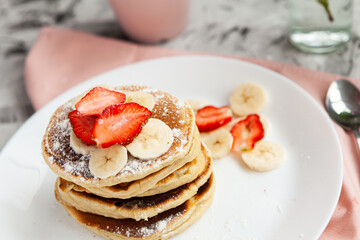A stack of pancakes with a pink tea mug and  fresh strawberries on a white plate on a light background. A small bouquet of lilacs in a glass vase stands next to the Breakfast.