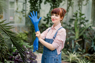 Attractive cute woman gardener in greenhouse, wearing blue rubber protective gloves, looking at camera, enjoying her work and smiling. Gardening, plants and watering concept