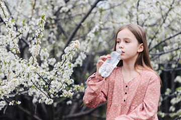 A little girl drinks clean fresh water from a plastic bottle near a flowering tree in the garden.