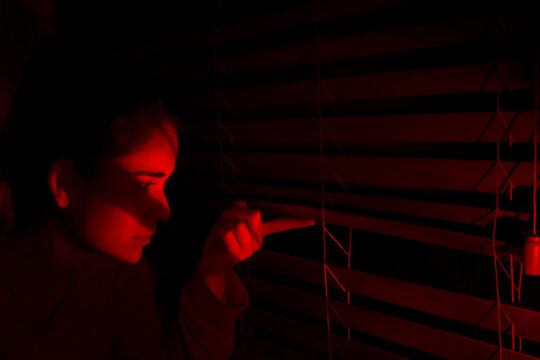 Curious young woman looking out the window. Girl spying through the blinds in the dark.