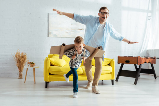 happy boy with carton plane wings, and cheerful father having fun while imitating flying