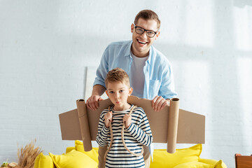 happy father and adorable boy with carton wings on back looking at camera