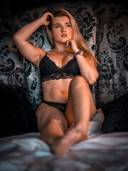 Boudoir session with a young blonde Caucasian woman on top of a bed with black lingerie, in a circular bed with a beautiful headboard of black flowers. Thoughtful posture