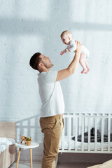 Fototapeta premium happy father holding adorable baby boy in outstretched hands near baby cot