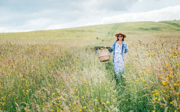Sincerely smiling young Woman dressed jeans jacket and light summer dress walking by the high green grass meadow with basket and wildflowers bouquet. Human and nature concept image.