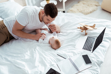 Obraz premium young father playing with baby boy near gadgets, notebook and toy plane on bed