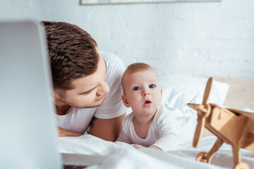 Obraz premium selective focus of young father looking at surprised baby boy while lying in bed near laptop and toy plane