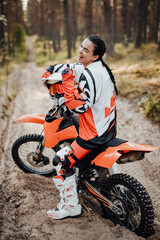 Happy brunette girl wearing motorcycle outfit sitting on her bike off-road in the woods