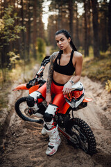 Cheeky tattooed racer girl wearing motocross outfit with semi naked torso sitting on her bike in the forest