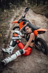 Tuinposter Beautiful female racer wearing motocross outfit with semi naked torso sitting on sand next to her bike in the forest © Fxquadro