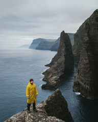 Traveler in a yellow raincoat on a cliff overlooking the rocks and ocean Faroe Islands. Tourism active life and adventures.
