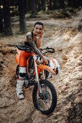 Fototapeta na wymiar Tattooed female racer wearing motocross outfit with semi naked torso sitting on her bike in the forest