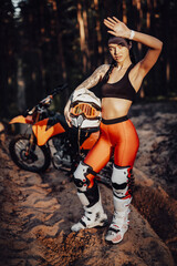 Beautiful brunette girl wearing motocross outfit with semi naked torso standing on a sand trail in the forest, holds his helmet and covers his eyes with his hand from the bright sun