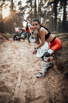 Young female racer wearing motocross outfit with semi naked torso sitting on a grass at sunset in the woods