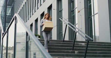 Caucasian upset businesswoman walking down the stairs outdoors with box of stuff as leaving business center. Female office worker lost her job. Unemployment rate growing at virus outbreak. Fired woman