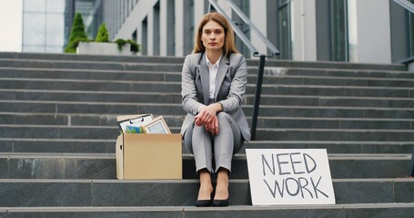 Portrait of Caucasian young workless woman siting on stairs at street with carton board Need Work and box with stuff. Female fired office worker in depression. Unemployment concept.