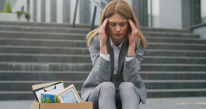 Portrait of Caucasian young workless woman siting on stairs at street with carton board Need Work and box with stuff. Female fired office worker in depression. Unemployment concept.