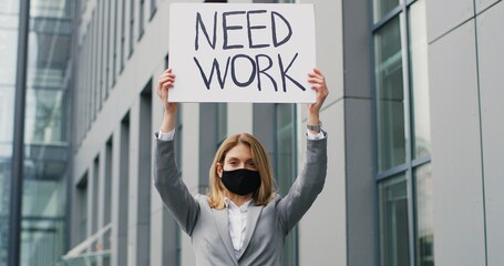 Portrait shot of Caucasian young woman demonstrating poster with words Need work. Female activist in mask showing board with looking for job announcement. Lonely protesting. Lockdown unemployment.