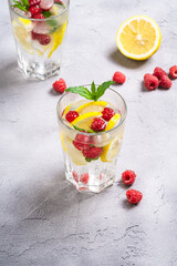 Fresh cold ice water with lemon, raspberry fruits and mint leaf in two faceted glass on stone concrete background, summer diet beverage, angle view