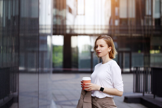 Business woman in beige suit, holding cup of coffee, walking outdoors on city street with skyscraper or building background, looking on window. Break at work, leisure time, enjoyment, shopping