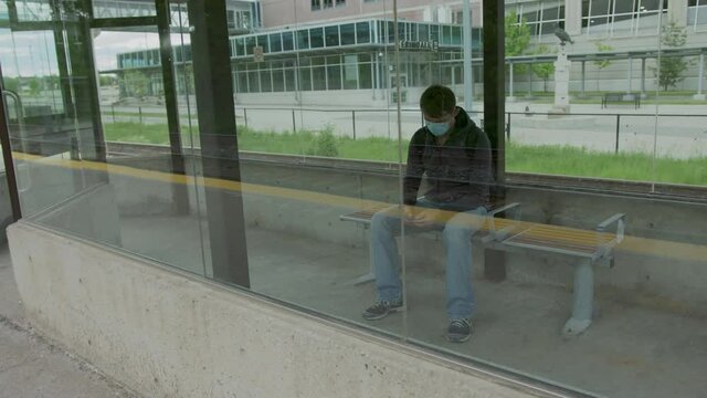 High quality 4k video of a young man who is waiting for a transportation in a glass bus stop and holding his cell phone in his hands. Man is texting on a station while waititng for a train 