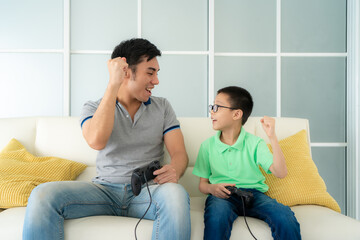 Asian family father and his son to playing video games with joysticks and arm raised to winning in game while sitting in sofa in living room at home, concept of family values.