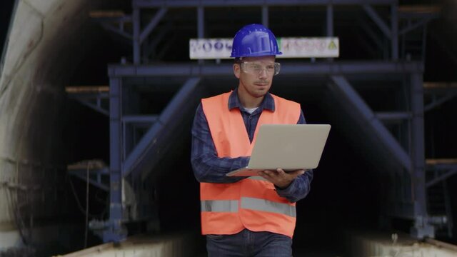 Construction engineer, architect or a supervisor wearing a protective equipment using a computer while exiting an underconstruction tunnel.