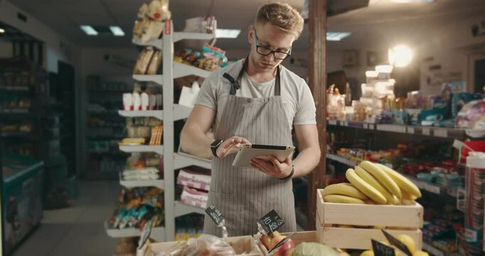 Bearded staff man in glasses and apron using digital tablet while standing near shelves with fruits and vegetables. Male worker doing accounting of products at supermarket.