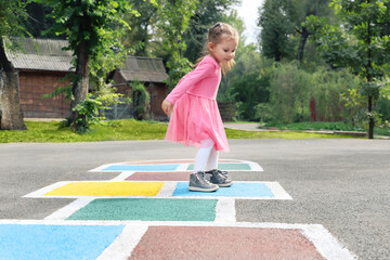 Little girl in a pink dress playing hopscotch on playground outdoors, children outdoor activities