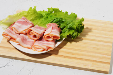 Rolled bacon slices are placed on a lettuce leaf on a plate. Bamboo cutting Board and texture table as background.