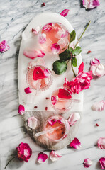 Summer refreshing cold beverage drink. Flat-lay of rose lemonade with ice in glasses and pink rose petals on oval serving board over grey marble table background, top view