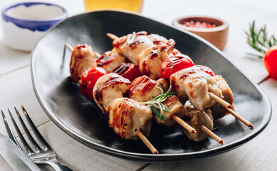 Grilled chicken skewers with cherry tomatoes on a black plate. White wooden table.