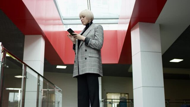 A close up slow motion shot of a blonde middle-aged business woman being in a hallway of an office building and reading through recieved messages on her phone