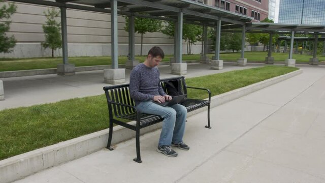 High quality 4k video of a young men who is sitting on a train station and working on his laptop which is holding on the lap. Young men sitting on a black metal banch on a station in the city. 