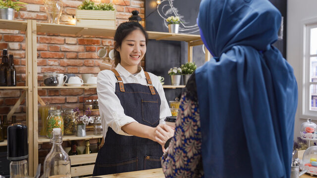 small business people and service concept. chinese woman bartender serving islam customer at coffee shop. friendly young girl waitress in apron giving disposable paper cup to muslim client in cafe.