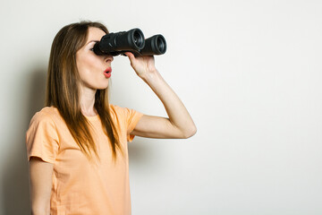 Young friendly girl in a T-shirt looks through binoculars on a light background