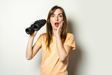 Young friendly girl with a surprised face in a T-shirt is holding binoculars on a light background. Banner. Emotional face