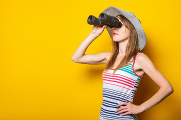 Young woman in hat and striped dress looks through binoculars on a yellow background.