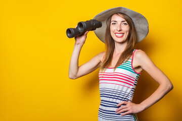 Friendly young smiling woman in a striped dress holds binoculars on a yellow background