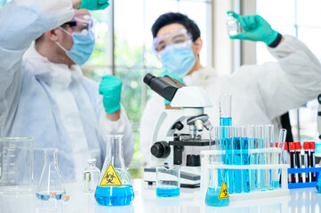 Lab equipment with blue liquid inside stand on the table with couple male scientist wearing protection suit holding vaccine for research at laboratory at the background.